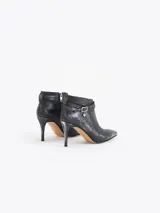 Bota corta de cuero vacuno color negro con acabado metalizado, taco fino mediano y punta fina. Presenta una tira decorativa con hebilla metálica y un dije en forma de corazón. Cuenta con cierre lateral para un calce cómodo y está completamente forrada en cuero.