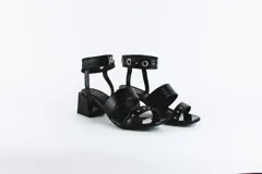 Sandalias negras de cuero con dos tiras horizontales sobre el pie y pulsera al tobillo con ojalillos y tachas. Taco cuadrado de 5 cm.