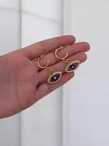 Caravanas tipo argolla con dije colgante en forma de ojo turco con pestañas doradas.