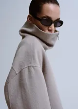 Chaqueta gris de diseño corto y holgado, con cuello alto envolvente, mangas anchas y puños ajustables con tiras y botones. Cuenta con bolsillos laterales y cierre frontal oculto.