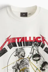 Remera de algodón color blanco crudo, corte oversized y manga corta. Presenta un estampado frontal grande con el logo de Metallica y la ilustración de Lady Justice, con el texto "AND JUSTICE FOR ALL" y "TOUR '83-'89".