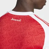 Camiseta de fútbol Adidas del Arsenal FC, modelo Primera Equipación 25/26, para hombre. Diseño principal rojo con estampado sutil de letras 'A' góticas, mangas y hombros blancos con tres franjas rojas. Cuello redondo.