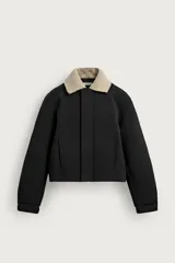 Cazadora boxy fit de tejido técnico negro con mezcla de algodón, de corte corto, manga larga con puños abotonados y cierre frontal con cremallera oculta. Cuenta con cuello solapa desmontable en tejido combinado color beige a contraste y bolsillos de vivo en la cadera.