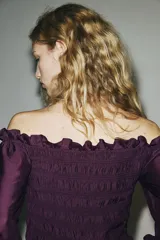 Blusa de color violeta con diseño de hombros descubiertos, cuerpo fruncido y mangas largas abullonadas con puños elásticos. Presenta detalles de volados en el escote, el bajo y los puños.