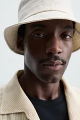 Sobrecamisa de hombre de corte relajado, color beige claro con finas rayas verticales en tono más oscuro. Presenta cuello bowling, manga larga con puño abotonado, bolsillos de parche en el pecho y en la cadera, y aberturas laterales en el bajo.