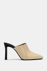 Zapato de tacón tipo mule en piel, de color beige con tacón fino y plantilla en contraste de color negro. Presenta escote redondo, costuras pespunteadas en los laterales y acabado en punta cuadrada.