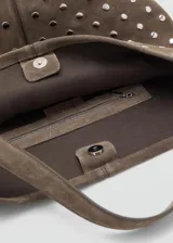 Bolso tipo hobo de cuero gamuzado color marrón, con diseño de tachas metálicas plateadas distribuidas por toda la superficie. Cuenta con una correa de hombro integrada y cierre superior.