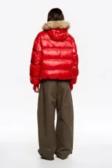 Chaqueta acolchada corta de tejido técnico brillante en color rojo, con cierre frontal de cremallera y botones de presión. Presenta capucha con borde de pelo sintético desmontable, bolsillos laterales con cremallera y puños de punto elástico en color rojo.