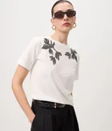 Remera blanca de manga corta y cuello redondo, confeccionada en algodón con bordados florales en tono oscuro aplicados en los hombros.