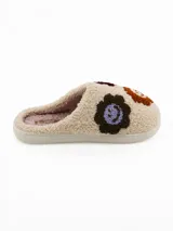 Pantuflas de estar por casa tipo mule, confeccionadas en tejido de peluche color crema. Presentan un diseño con apliques bordados de caritas sonrientes en tonos marrones, bordó y lila. Cuentan con una suela plana de goma y forro interior suave.