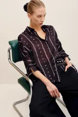 Camisa de seda negra con estampado geométrico de líneas verticales con rombos pequeños en tonos claros, cuello camisero, mangas largas con tajitos en los puños y cierre frontal con botones.