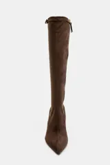 Bota alta de caña media en color marrón, confeccionada en material tipo gamuza. Presenta un diseño de punta fina, tacón alto y fino, y una tira decorativa con hebilla metálica en la parte superior de la caña. Incluye cierre lateral mediante cremallera.