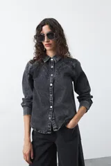 Camisa de jean color azul claro con tachas metálicas en el canesú y a lo largo de las costuras frontales. Tiene mangas largas con puños abullonados y cierre frontal con botones.