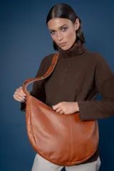 Bolso tipo bandolera de gamuza color marrón, con correa ancha ajustable decorada con tachas metálicas y cierre superior.