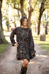 Vestido corto de lino con estampado floral en tonos tierra y verdes. Presenta cuello redondo, mangas largas amplias con puños terminados en volados y falda con volado en el ruedo. Incluye un lazo desmontable para ajustar la cintura.