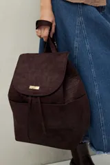 Mochila de símil gamuza color negro con textura tipo reptil. Presenta un diseño minimalista con solapa frontal, cierre de cordón ajustable y bolsillos laterales. Incluye un detalle metálico rectangular en la solapa.