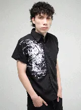 Camisa de manga corta color negro, con corte regular y cierre frontal de botones. Presenta un estampado gráfico en blanco en el lado derecho del pecho con motivos artísticos y tipografía gótica.