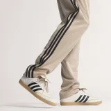 Pantalón de jogging Adidas de corte regular en color beige, con las tres tiras características en negro a lo largo de los laterales y el logo del trifolio bordado en la parte frontal.