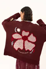 Saco de punto color vino con diseño holgado, cuello redondo y mangas largas. Presenta un gran bordado central en la espalda con forma de flor en tonos crema y rojo, acompañado de un pequeño bordado de sol y una frase bordada en la parte inferior.