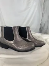 Botín tipo Chelsea de cuero metalizado en tono bronce, con paneles elásticos laterales en color beige. Presenta detalles de costuras decorativas tipo brogue en la puntera y laterales, con suela de goma antideslizante y taco bajo foliado.