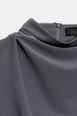 Vestido midi negro con cuello subido y diseño sin mangas (sisa). Presenta un elegante detalle de tejido drapeado en el lateral y cierre trasero con cremallera oculta.