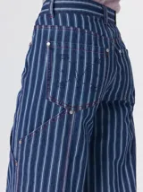 Pantalón de tiro medio y pierna ancha confeccionado en denim de algodón grueso. Presenta un diseño a rayas verticales en tonos azul y celeste, con costuras en contraste color rojo, bolsillos laterales tipo cargo y remaches metálicos.