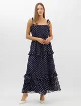 Vestido largo azul marino con estampado de lunares blancos, confeccionado en gasa fluida. Cuenta con tirantes finos ajustables con lazos en los hombros, escote con volados, corte en la cintura con lazo ajustable, y falda larga con múltiples capas y volados horizontales.