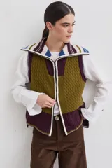 Chaleco de lana con cierre frontal completo y cuello alto. Presenta un diseño de bloques de color en tonos marrón chocolate y celeste, combinando secciones de tejido trenzado con secciones de tejido acanalado. Posee un corte asimétrico, siendo más largo en la parte posterior, y aberturas laterales en el ruedo.