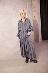 Camisa de lino con diseño a rayas verticales en tonos gris y azul. Presenta cuello solapa, cierre frontal con botones, mangas largas abullonadas con puños elásticos y un corte holgado que se cruza en la cintura.