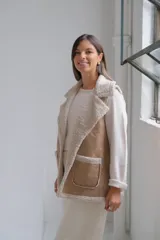 Chaleco de gamuza color beige con interior y detalles de corderito color crudo. Presenta solapas amplias, bolsillos frontales aplicados y diseño sin mangas.