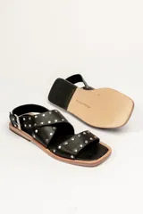 Sandalias bajas de cuero negro con tachas plateadas, tiras cruzadas y hebilla al tobillo.