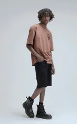 Remera manga corta color marrón (mocca) con un gran estampado trasero en negro que presenta tres máscaras o calaveras estilizadas con una corona en la parte superior y gotas o lágrimas cayendo.