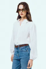 Camisa blanca de manga larga con sutil patrón de rayas verticales, cuello camisero, cierre frontal con botones y puños elásticos.