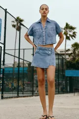 Camisa corta de denim en color azul claro, con cuello solapa, escote en V y manga corta. Presenta bolsillos delanteros de parche con solapa y cierre frontal de botones metálicos.