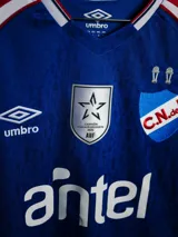 Camiseta de arquero Umbro del Club Nacional de Football, temporada 2026. Diseño en color azul con detalles en rojo y blanco en los hombros, cuello en V y estampado sutil en la tela. Incluye logo de Umbro, escudo del club, logo de la AUF y sponsor Antel en el frente.