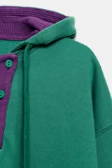 Sudadera con capucha Fruit of the Loom en colaboración con Zara, de color verde con detalles en morado en el interior de la capucha, la cartera de botones y los bordes del bolsillo frontal tipo canguro. Presenta mangas largas, cordones ajustables y un parche con el logo en el centro.