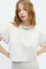 Blusa blanca de manga corta con cuello redondo y bordado de lentejuelas y mostacillas plateadas en el cuello y las mangas.