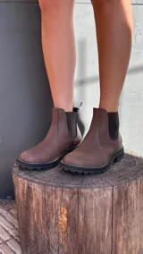 Bota tipo Chelsea de cuero marrón con textura granulada, paneles laterales elásticos en color negro y suela track dentada con costura blanca visible.