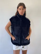 Chaleco de peluche negro con cuello alto, cierre frontal, bolsillos delanteros y detalles decorativos en los hombros.