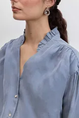 Camisa de satén color azul grisáceo, con cuello mao con detalle de volados, cierre frontal con botones y mangas largas con puños abotonados.