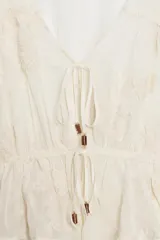 Blusa de escote en pico y manga larga con puños elásticos. Presenta un diseño semitransparente con bordados florales a tono, cintura elástica y cierre frontal mediante lazos con abalorios decorativos.