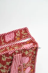 Bolso tipo tote acolchado con estampado floral en tonos rojizos, rosados y verdes. Presenta asas de mano con diseño de rayas verticales en color rojo y blanco, y ribetes a juego en los bordes.