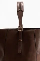 Cartera shopper de diseño slouchy en color marrón, con correa ajustable en la parte superior y detalles de tiras colgantes en los laterales con hebillas metálicas.