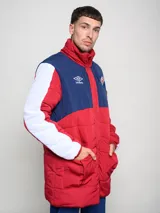 Parka larga de abrigo Umbro del Club Nacional de Football, con diseño tricolor en rojo, azul y blanco. Presenta cuello alto, cierre frontal con solapa, bolsillos con cierre en el pecho y logo de la marca junto al escudo del club.