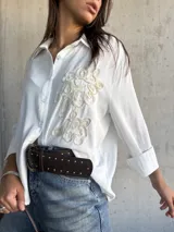 Camisa blanca de corte holgado con cuello clásico, cierre frontal de botones y detalle de bordado en relieve en el frente. Posee mangas largas con puños doblados.