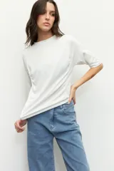 Remera blanca de textura sutil, con cuello a la base, mangas tres cuartos y detalle de fruncidos laterales que aportan movimiento y caída a la prenda.