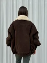 Campera de gamuza color marrón con cuello de corderito sintético en color crema. Presenta un diseño oversize con cierre frontal, solapas con broches metálicos y una tira ajustable en el ruedo.