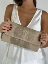 Cartera de mano tipo sobre color beige con textura símil piel de cocodrilo, solapa con cierre magnético y cadena dorada desmontable para usar al hombro.