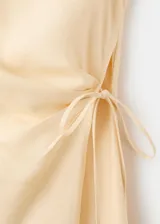 Vestido midi color beige, de cuello redondo y sin mangas, con diseño cruzado en la falda y lazo ajustable en la cintura.
