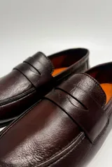 Mocasín de cuero color café, diseño clásico de estilo penny loafer con suela plana y costuras visibles.